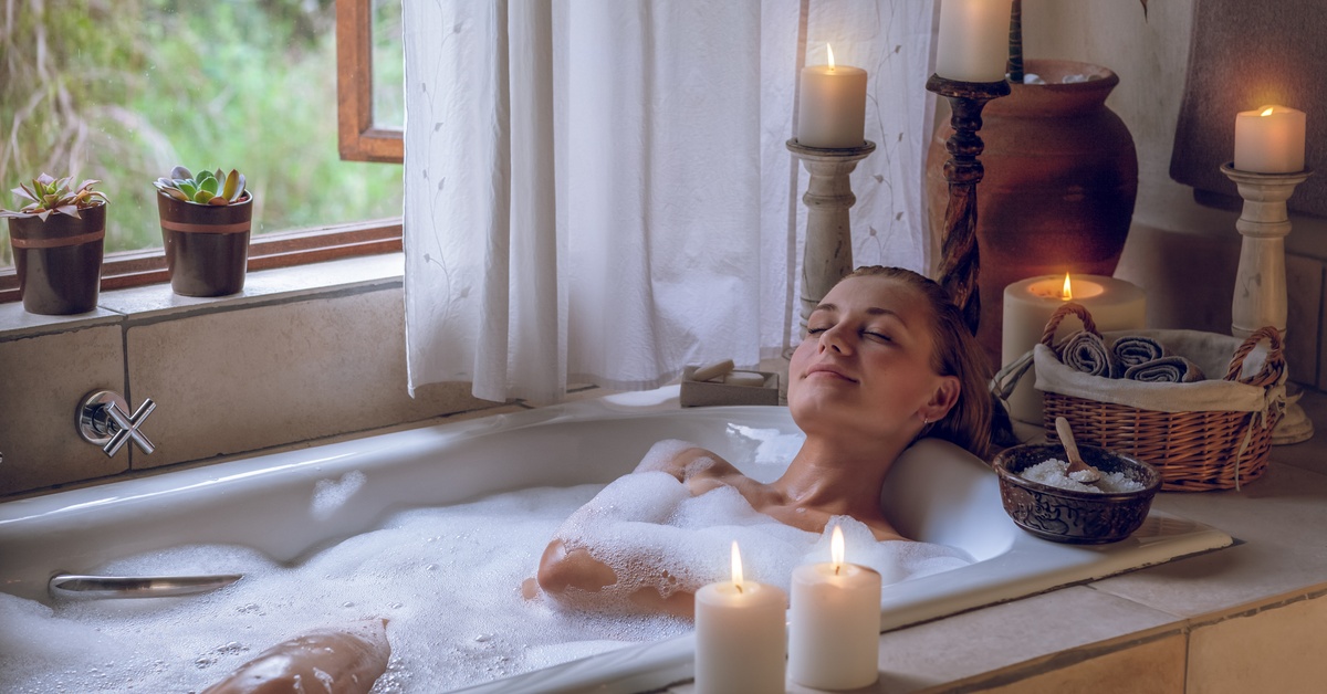 A woman closes her eyes as she enjoys a bubble bath. Around her are calming candles, plants, and an open window.
