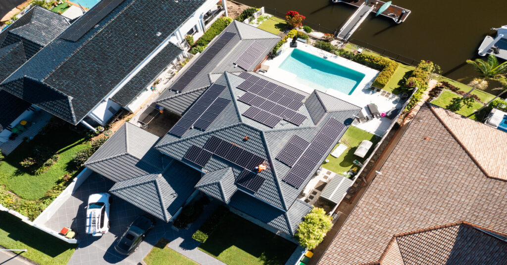 A technician is working on solar panels installed on a luxury waterfront home under a clear, sunny sky.