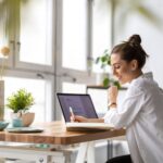 The Ideal Home Office Setup for the Remote Worker A woman sits at a wooden desk in front of large, picturesque windows. A laptop is open, and she's writing in a notebook.