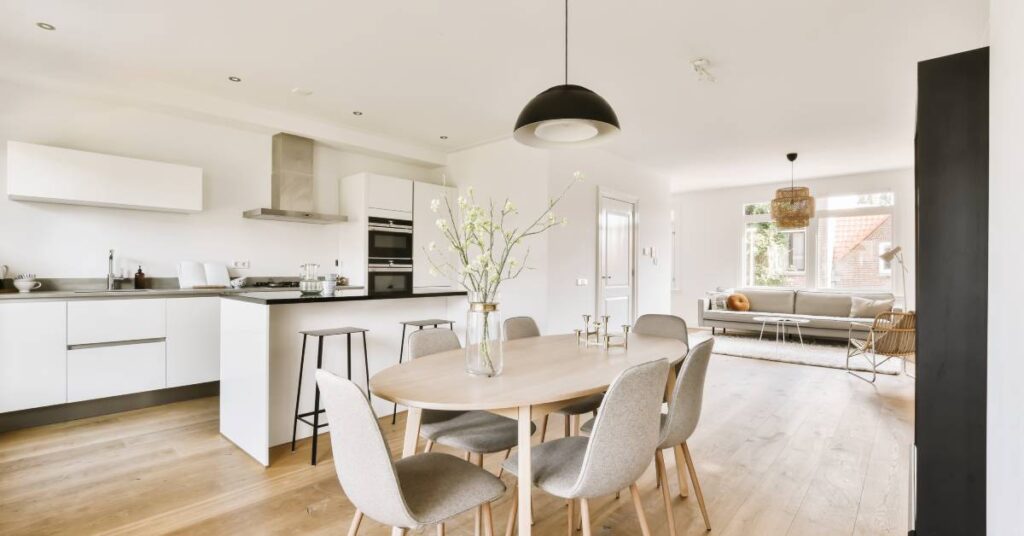 A dining table with six gray chairs stands in front of a kitchen with white cabinets and black countertops.