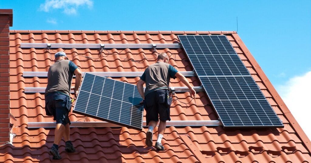 Two men wearing brown T-shirts and blue shorts are installing a solar panel on a stucco roof on a sunny day.