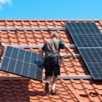 Two men wearing brown T-shirts and blue shorts are installing a solar panel on a stucco roof on a sunny day.
