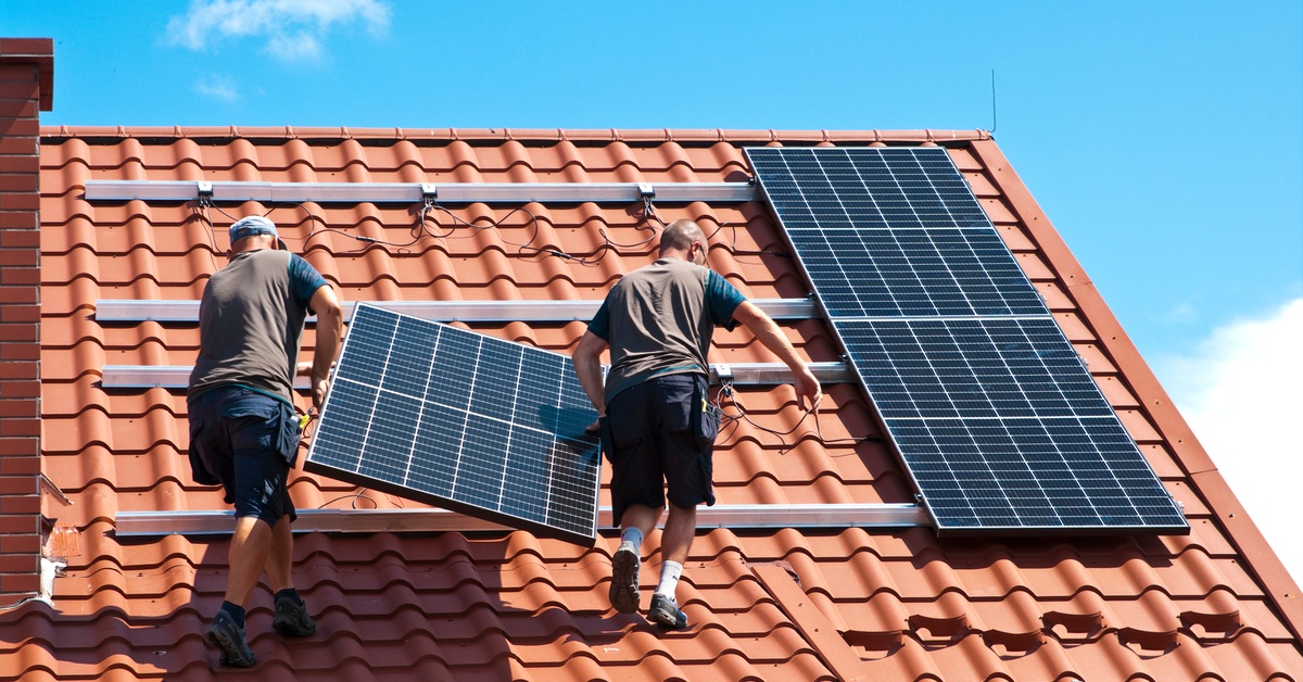 Two men wearing brown T-shirts and blue shorts are installing a solar panel on a stucco roof on a sunny day.