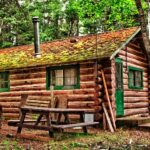 5 Essential Construction Projects for Log Home Owners A rustic log cabin nestled in the woods surrounded by trees with a metal chimney and a picnic table next to it.