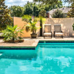 Exterior Additions That Increase Your Home’s Property Value Closeup of a luxurious backyard pool with bright turquoise water next to a concrete patio with chairs and plants.