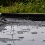 Common Problems Related to Flat Roofing Systems A flat black roof with rain pouring over it, pooling into a puddle on top of the house with trees in the background.