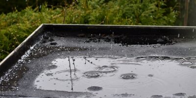A flat black roof with rain pouring over it, pooling into a puddle on top of the house with trees in the background.