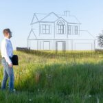 5 Reasons To Purchase Land Before Building Your Dream House A man is standing in the middle of a field, imagining a newly built, two-story home with a tree planted next to it.