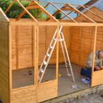 An older man wearing a jacket and jeans kneeling down with a power tool while working on a wooden building.