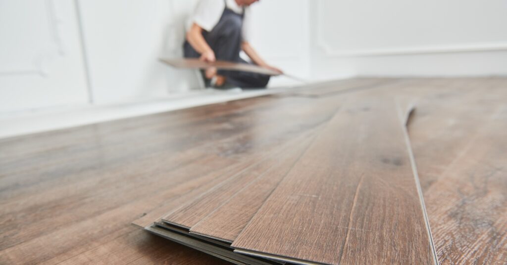 A stack of luxury vinyl flooring planks is in focus in the foreground, and a worker is installing the floor in the background.