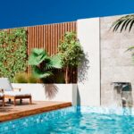 4 Amazing Hardscaping Design Options for Luxury Homes A modern outdoor pool with turquoise-colored water, two lounge chairs on a wooden deck, and green plants nearby.