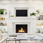 What To Know Before Buying a Home With a Fireplace A luxurious home living room with a large fireplace in the center surrounded by white accents, decor, and furniture.