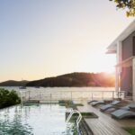4 Luxury Upgrades That Will Elevate Your Beach House The sun sets on a modern, luxury beach house with a large rectangular pool. Sailboats float on the ocean in the background.