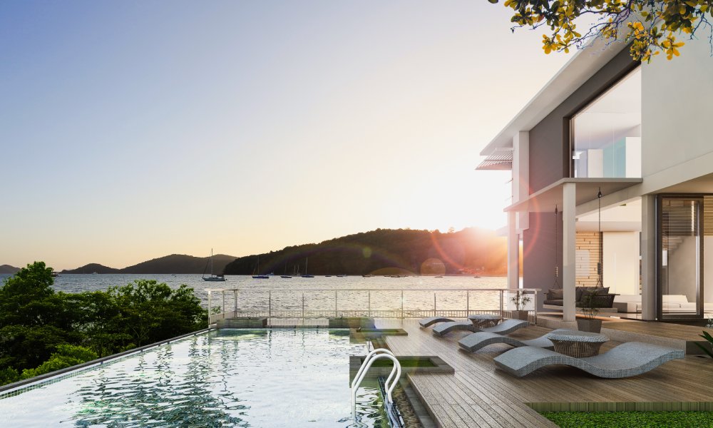 The sun sets on a modern, luxury beach house with a large rectangular pool. Sailboats float on the ocean in the background.