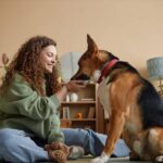 A smiling young woman sits on the living room floor feeding her dog a treat. A mirror and a bookshelf are in the background.
