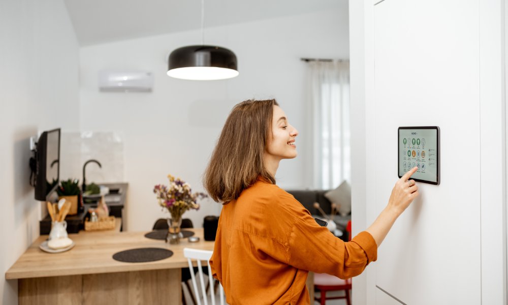 A woman wearing a burnt orange blouse uses a smart home thermometer to control the house's temperature.