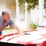 Why You Should Wash Your Car More in the Summer An older man is washing his vintage red luxury sports car with white stripes. He's applying wax to the hood.