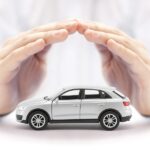 6 Common Factors That Impact Your Car Insurance Rate A close-up view shows a small, white toy SUV sitting on a white surface with a pair of hands forming a cover over it.