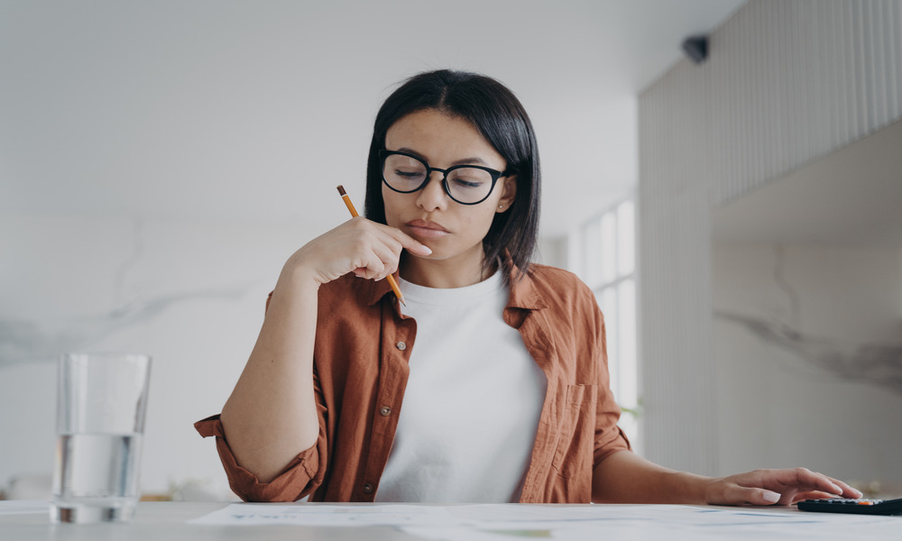 A woman wearing glasses sits at a table while looking at some paperwork and holding a pencil under her chin.