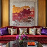 A red velvet sofa with purple and patterned pillows sits below abstract art and gold sconces on the wall.