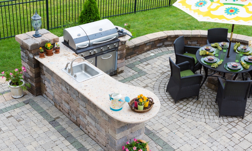 A patio in the backyard of a home with a grill, countertop, and table for dining in front of a black fence.